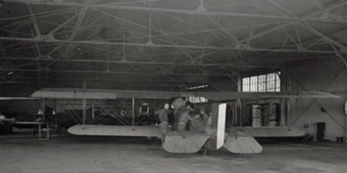 A vintage aircraft inside a hangar, with two people working on it. The space features large windows and metal beams.