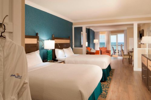 A bright hotel room with two beds, a balcony view, and coastal decor, featuring a robe hanging on the door.