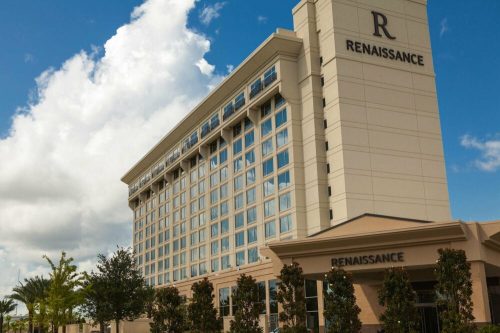 A modern Renaissance hotel building with large windows, surrounded by palm trees and a blue sky with clouds.
