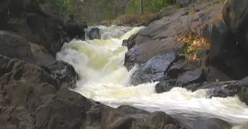 A rushing river flows over dark rocks, surrounded by trees and autumn foliage.