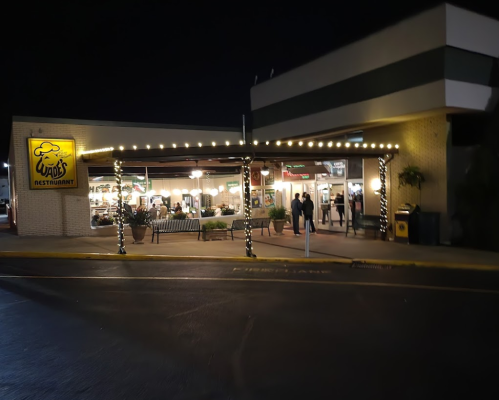 A brightly lit restaurant exterior at night, featuring outdoor seating and festive lights.