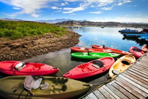 Colorful kayaks lined up on a shore by a calm lake, with mountains and a blue sky in the background.