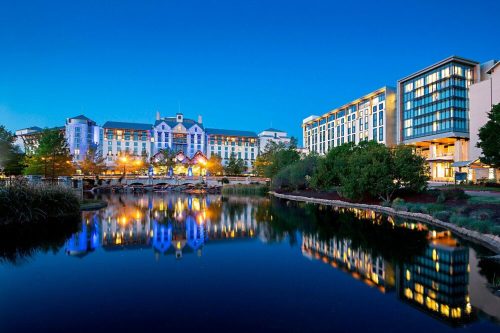 A serene hotel scene at dusk, reflecting in a calm lake with colorful lights and lush greenery surrounding the area.