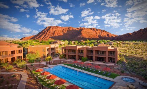 A scenic view of a resort with a pool, surrounded by red rock mountains and blue skies with scattered clouds.