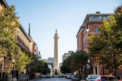 A tall monument stands at the end of a tree-lined street, flanked by historic buildings under a clear blue sky.