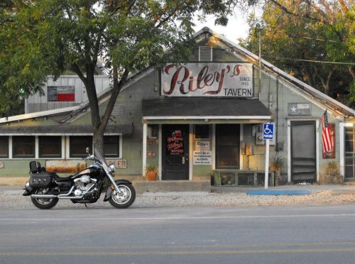 A motorcycle parked in front of Riley's Tavern, a rustic building with a sign and trees lining the street.