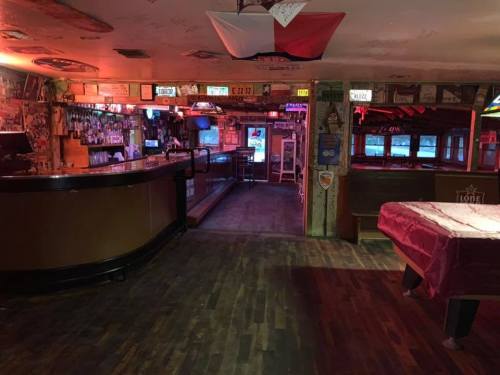 A dimly lit bar interior featuring a wooden counter, pool table, and various signs on the walls.