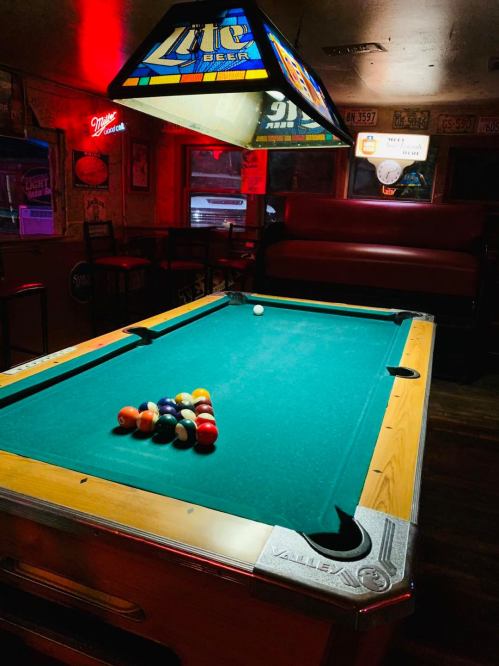 A dimly lit pool table with a triangle of balls, under a colorful beer sign in a cozy bar setting.