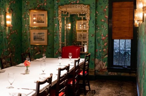A dining room with a green floral wallpaper, a long table set for dinner, and red chairs, illuminated by soft lighting.