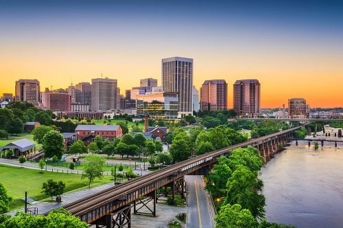 A vibrant city skyline at sunset, featuring buildings, greenery, and a river with a train track in the foreground.