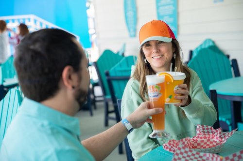 A woman in a colorful cap smiles while toasting drinks with a man at a bright outdoor café.