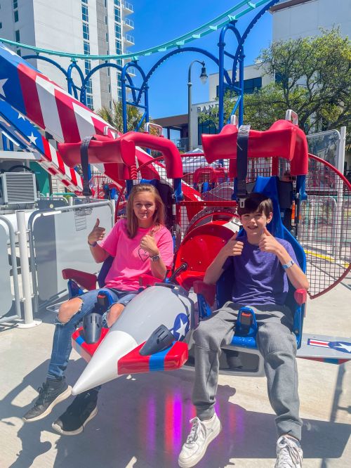 Two kids sit in a colorful amusement park ride, giving thumbs up, with a patriotic theme in the background.