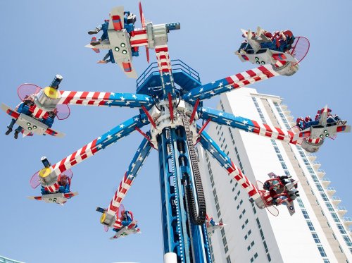 A colorful amusement park ride with seats shaped like planes, spinning against a clear blue sky and tall buildings.
