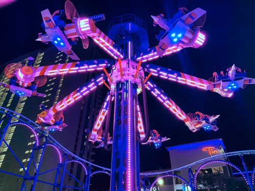 A colorful amusement ride with spinning seats, illuminated by vibrant lights at night, set against a city backdrop.