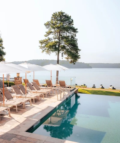 A serene poolside view with lounge chairs, umbrellas, and a lake in the background, surrounded by trees.