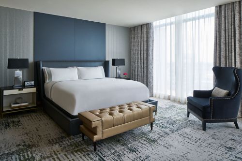 Modern hotel bedroom featuring a large bed, stylish armchair, and large windows with sheer curtains.