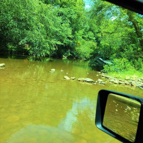 A serene river scene surrounded by lush greenery, viewed from a vehicle's side mirror.