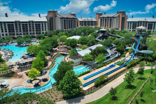 Aerial view of a resort with a large water park, featuring slides, pools, and lush greenery under a blue sky.