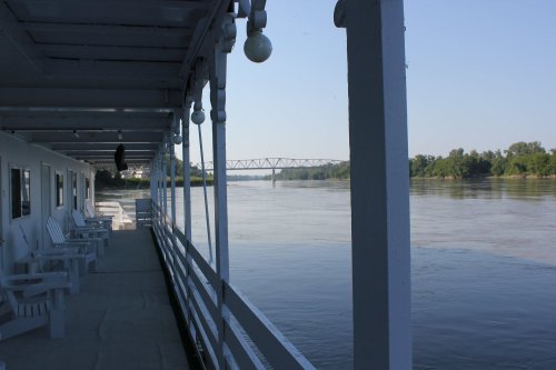 View from a boat deck overlooking a calm river with a bridge in the distance and white chairs lined up along the railing.