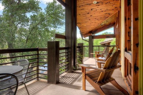 A wooden balcony with two chairs overlooks a lush green landscape and trees under a bright blue sky.