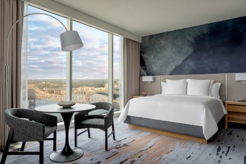 Modern hotel room with a large window view, featuring a bed, table, and chairs, overlooking a cityscape.