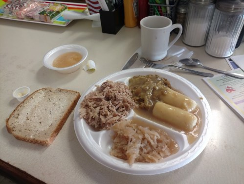 A plate with shredded meat, dumplings, sauerkraut, a slice of bread, a cup of applesauce, and a coffee mug on a table.