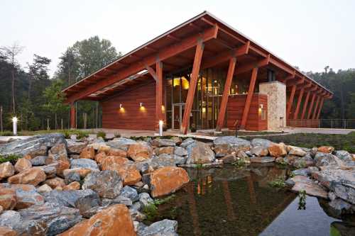 Modern wooden building with a sloped roof, surrounded by rocks and a small pond, set in a natural landscape.