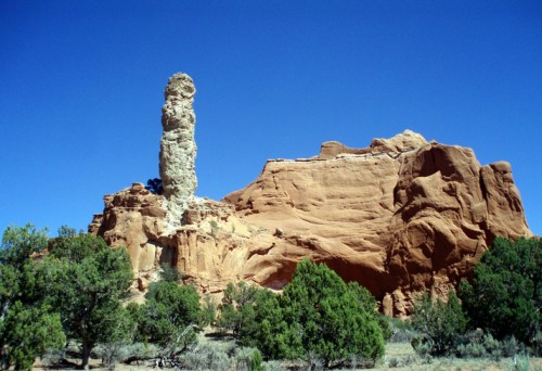 A tall rock formation stands prominently against a clear blue sky, surrounded by desert vegetation and red rock cliffs.
