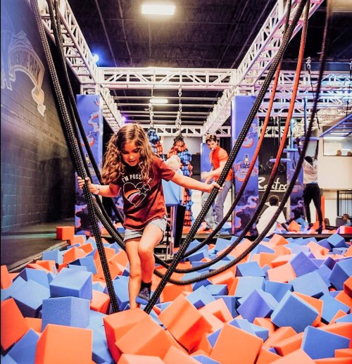 A girl navigates through foam blocks while balancing on ropes in a vibrant indoor adventure park.