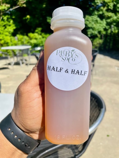A hand holding a bottle labeled "Ruby's Half & Half," with a clear liquid inside, outdoors in a sunny setting.