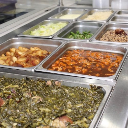 A variety of dishes in metal trays, including greens, chicken, potatoes, and vegetables, arranged in a cafeteria setting.