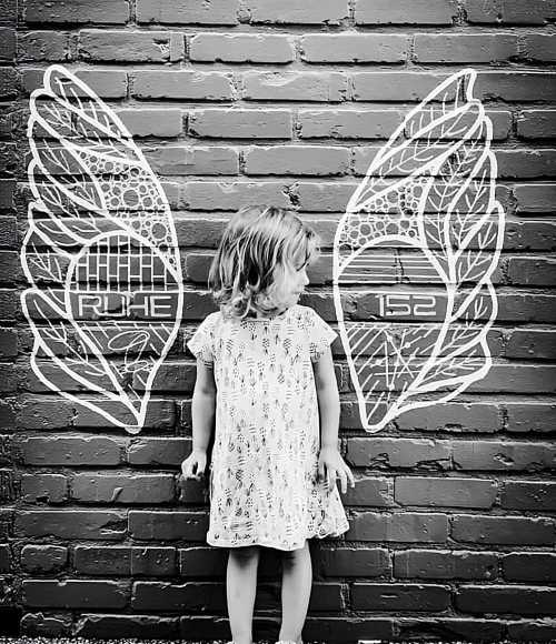 A child stands in front of a brick wall with white wing murals, looking to the side in a playful pose.