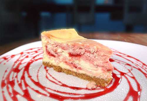 A slice of strawberry cheesecake on a white plate, drizzled with raspberry sauce and dusted with powdered sugar.