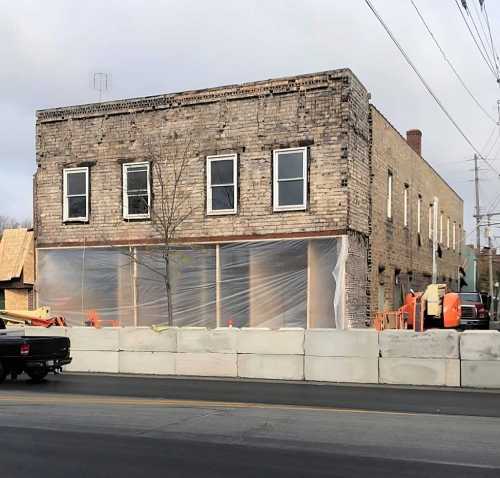 A brick building under renovation, covered with plastic sheeting, with construction equipment nearby and a truck in the foreground.