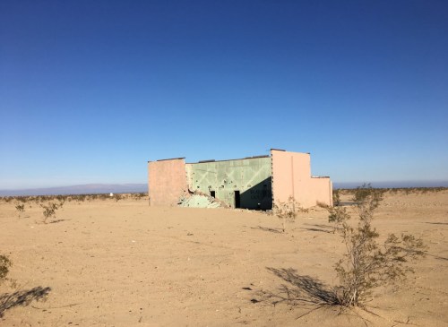 A weathered, abandoned building stands alone in a vast desert landscape under a clear blue sky.