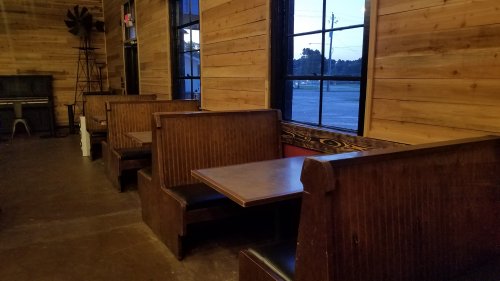 Wooden booths with tables line a rustic interior, featuring large windows and warm wooden walls.