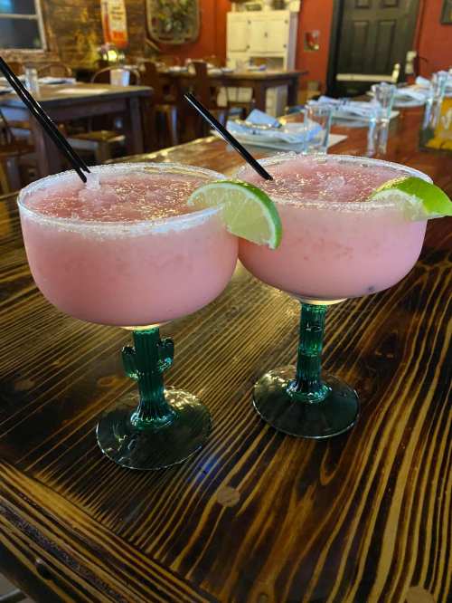 Two pink margaritas with lime wedges, served in green glasses, on a wooden table in a restaurant setting.