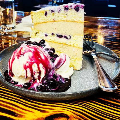 A slice of layered cake with blueberries, served with a scoop of vanilla ice cream and blueberry sauce on a gray plate.