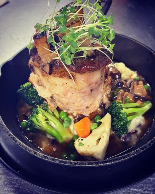 A beautifully plated dish featuring a roasted meat cut topped with microgreens, surrounded by colorful vegetables in sauce.