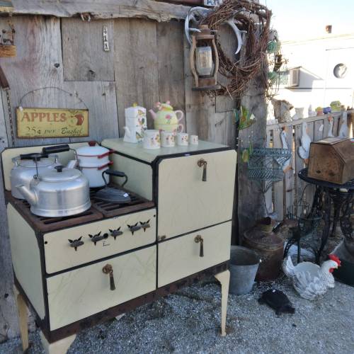 A vintage kitchen setup with an old stove, teapot, and decorative mugs against a rustic wooden backdrop.