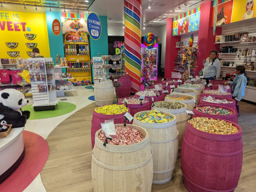 A colorful candy store filled with barrels of sweets, vibrant decor, and a panda plush toy in the corner.