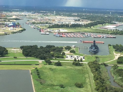 Aerial view of a river with a ship, docks, and green parks under a cloudy sky.