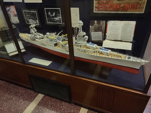 A detailed model of a battleship displayed in a glass case, surrounded by historical photographs and documents.