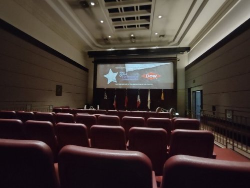 Empty auditorium with red seats, a large screen displaying a logo, and flags in the background.