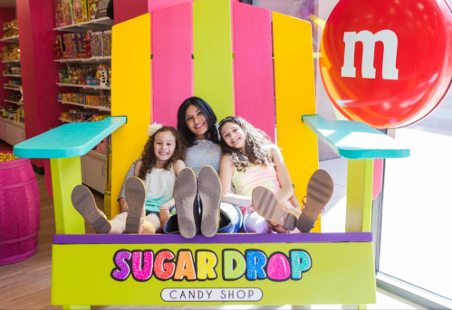 A colorful oversized chair at a candy shop with two girls and a woman sitting together, all smiling.