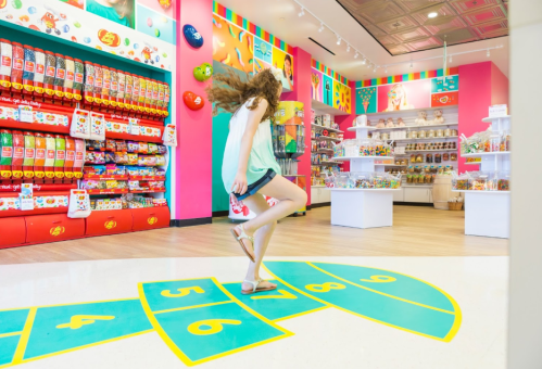 A girl jumps on a colorful hopscotch floor in a vibrant candy store filled with shelves of sweets.