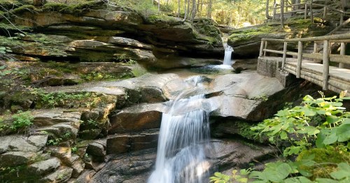 A serene waterfall cascades over rocks, surrounded by lush greenery and a wooden walkway in a tranquil forest setting.