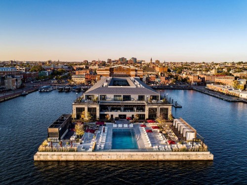 Aerial view of a modern waterfront building with a pool, surrounded by cityscape and water. Clear blue sky above.