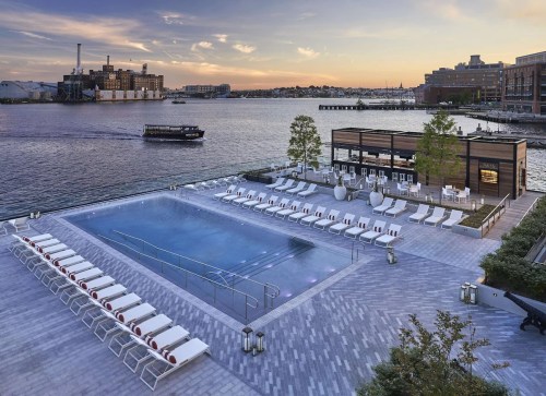 Rooftop pool with lounge chairs overlooking a waterfront at sunset, with a boat in the distance.