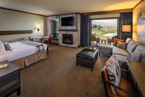 Cozy hotel room with a king bed, seating area, fireplace, and large windows overlooking a scenic landscape.